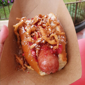 50th Celebration hotdog - Runder Hot Dog gegarneerd met Aardbei-Bacon Jam, Funnel Cake stukjes en Poedersuiker.